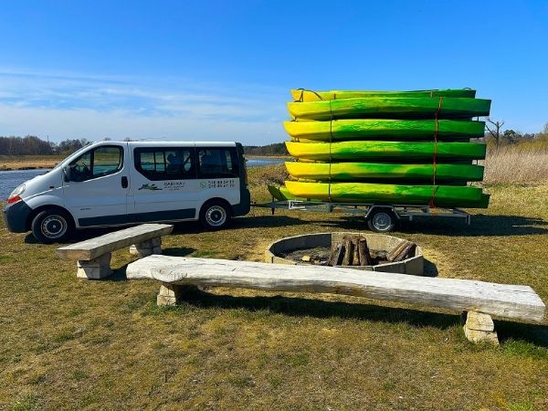 Biały samochód dostawczy z przyczepą, na której ułożony jest wysoki stos zielono-żółtych kajaków. Zespół pojazdów stoi na trawiastym terenie obok miejsca na ognisko z drewnianymi ławami, w pobliżu brzegu wody pod bezchmurnym niebem.