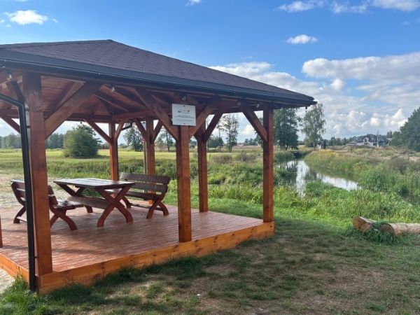 Drewniana, zadaszona altana z ławami i stołem, ustawiona na murowanym podłożu nad brzegiem rzeki. W tle rozpościera się zielony krajobraz z łąkami i drzewami pod błękitnym niebem z białymi chmurami.